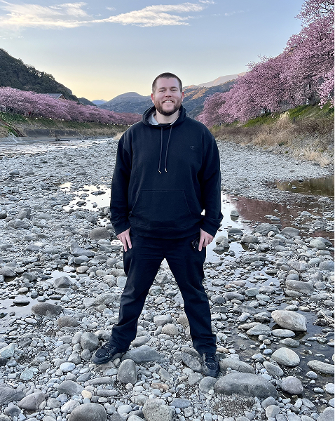 Chris standing on a stony riverbed under cherry blossoms at sunset